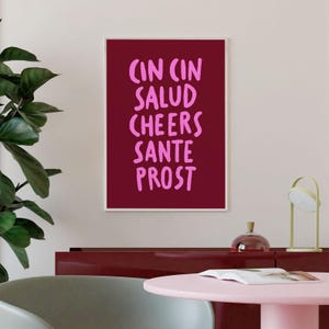 Peut inclure: Une affiche encadr&eacute;e avec un fond bordeaux et du texte rose. Le texte comprend les mots "CIN CIN, SALUD, CHEERS, SANTE, PROST". L'affiche est affich&eacute;e sur un mur au-dessus d'un meuble rouge fonc&eacute; et d'une table rose.