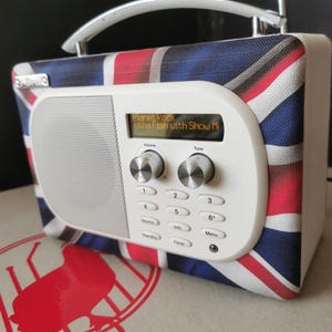 May include: A DAB radio with a Union Jack design. The white front panel has a digital display showing radio information. It has volume and tuning knobs, and a silver handle. The radio is a portable design.