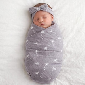 Könnte beinhalten: Ein neugeborenes Baby, eingewickelt in eine graue Decke mit einem weißen Pusteblumenmuster. Das Baby trägt ein passendes graues Stirnband mit einer Schleife. Das Baby schläft friedlich auf einer weißen Oberfläche.