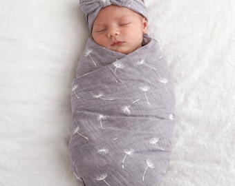 May include: A newborn baby wrapped in a gray swaddle blanket with a white dandelion pattern. The baby wears a matching gray headband with a bow. The baby is sleeping peacefully on a white surface.