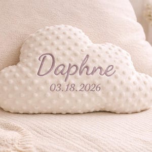 May include: A white cloud-shaped cushion with a textured surface. The name "Daphne" and the date "03.18.2026" are embroidered in a soft purple thread. The cushion is placed on a cream-coloured surface.