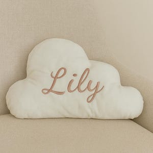 May include: A cloud-shaped cushion in an off-white colour featuring the name Lily embroidered in a cursive script using a dusty rose thread. The cushion has a soft, plush texture and is centred with the name clearly visible on the front surface.