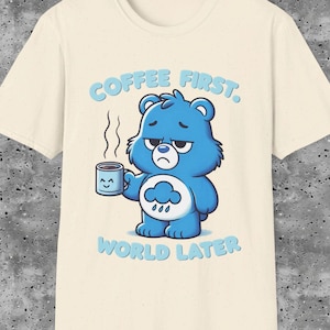Puede incluir: Una camiseta beige claro con un oso de dibujos animados azul sosteniendo una taza de café. El texto "COFFEE FIRST. WORLD LATER." está impreso en la camiseta.