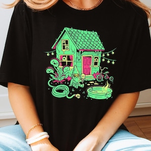 Może przedstawiać: Czarny t-shirt z nadrukiem graficznym przedstawiającym neonowo-zielony dom z różowymi drzwiami, krasnala ogrodowego trzymającego rożek lodowy, rower, zwinięty wąż ogrodowy, małe palenisko oraz rośliny mięsożerne w odcieniach zieleni i różu.