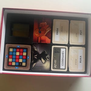 May include: A red box with a black and white game board, a red and blue grid, and several cards with text and images. The cards include the words "CODENAMES", "RUKA", "PUT", "PERO", and "#2 HR".