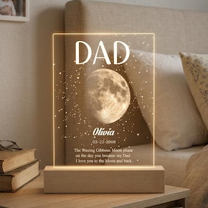 May include: A personalised night light featuring the word "DAD", a moon graphic, and the name "Olivia". The light sits on a wooden base and includes a message about the moon phase.