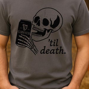 May include: Dark gray t-shirt featuring a black graphic of a skull holding a Guinness can with a skeleton hand. The text "'til death." is below the skull. The shirt is a crew neck and has short sleeves. Pumpkins and fall foliage are in the background.