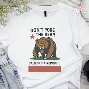 May include: White t-shirt with a graphic design featuring a brown bear with an angry expression, a red star, and the text "DON'T POKE THE BEAR" above the bear. Below the bear is the text "CALIFORNIA REPUBLIC" and a red rectangle.