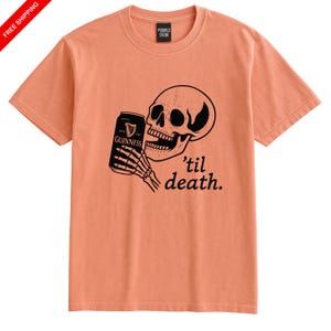 May include: Peach-colored t-shirt featuring a black graphic of a skull holding a Guinness can. The text "'til death" is below the skull. The shirt has a crew neck and short sleeves.