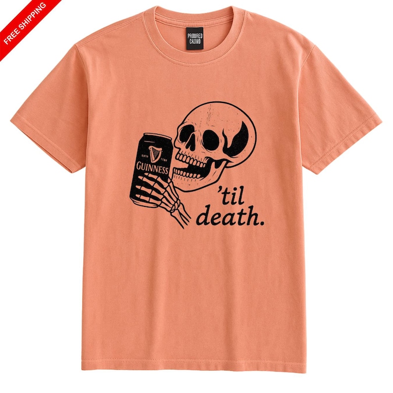 May include: Peach-colored t-shirt featuring a black graphic of a skull holding a Guinness can. The text "'til death" is below the skull. The shirt has a crew neck and short sleeves.