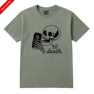 May include: Sage green t-shirt featuring a black graphic of a skull holding a Guinness can, with the phrase "'til death." printed below. The shirt has a crew neck and short sleeves.