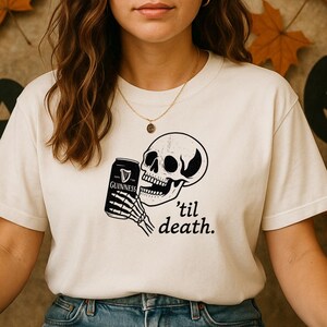 May include: Cream-colored t-shirt featuring a black and white graphic of a skull holding a Guinness can. The phrase "'til death." is printed below the skull. The shirt is a casual style.