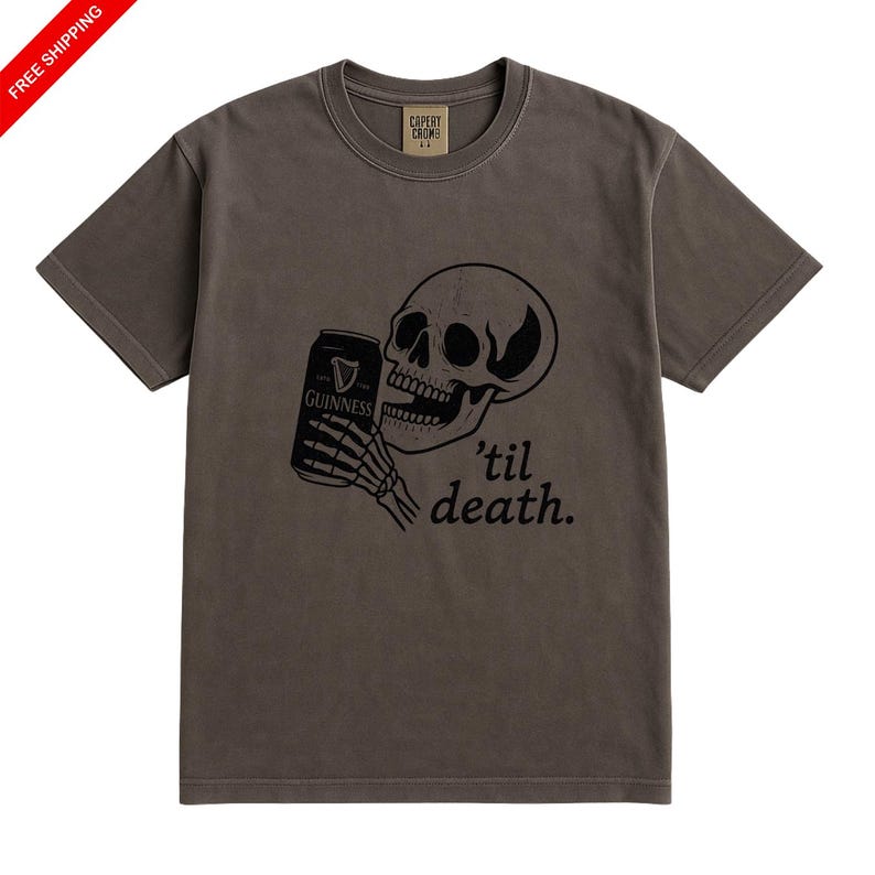 May include: A brown t-shirt features a black graphic of a skull holding a Guinness can with the text "'til death." The shirt has a crew neck and short sleeves. The brand name "Capery Crows" is printed on the upper back.