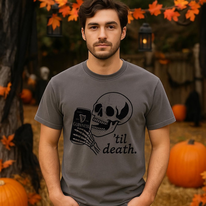 May include: Dark gray t-shirt featuring a black graphic of a skull holding a Guinness can with a skeleton hand. The text "'til death." is below the skull. The shirt is a crew neck and has short sleeves. Pumpkins and fall foliage are in the background.