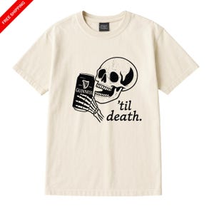 May include: Cream-colored t-shirt featuring a black and white graphic of a skull holding a Guinness can. The phrase "'til death" is printed below the skull. The shirt has a crew neck and short sleeves.