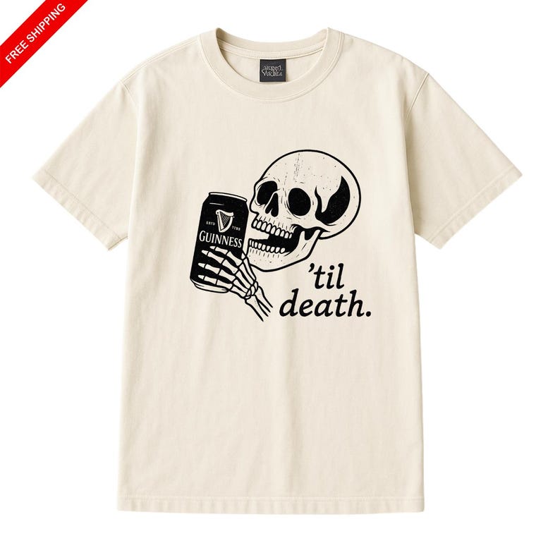 May include: Cream-colored t-shirt featuring a black and white graphic of a skull holding a Guinness can. The phrase "'til death" is printed below the skull. The shirt has a crew neck and short sleeves.