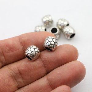May include: Silver metal beads with a textured, geometric design. The beads have a hole in the center for stringing.