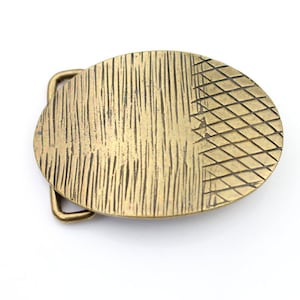 Op de afbeelding: Ovale messing riemgesp met een getextureerd oppervlak. De gesp heeft een verticaal lijnenpatroon aan de linkerkant en een ruitpatroon aan de rechterkant. Het heeft een rechthoekige lus voor de riem.