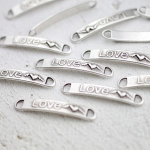 May include: A close-up of several silver metal charms with the word "Love" and a heart with wings engraved on them. The charms are scattered on a white surface.