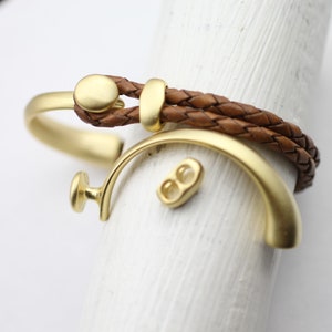May include: Two gold-toned bracelets, one a braided leather bracelet with a gold clasp and the other a simple bangle with a rounded end. A small gold connector is also visible.