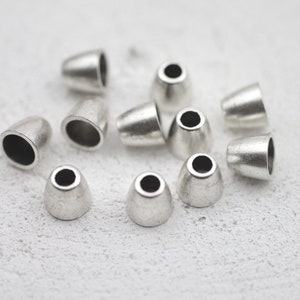 May include: A group of silver metal cord ends, also known as cord tips, are scattered on a white surface. These metal cord ends are used to finish the ends of cords and prevent fraying.