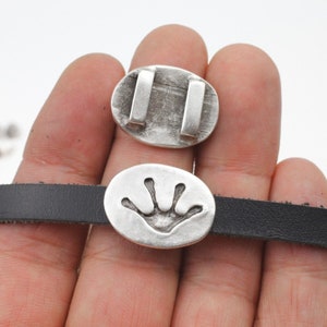 Könnte beinhalten: Zwei ovale Charms aus silberfarbenem Metall. Ein Charm hat ein erhabenes Design einer Hand mit vier Fingern. Der andere Charm hat zwei parallele Linien.