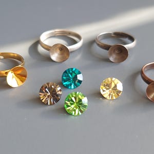 May include: An assortment of rings with settings and colorful gemstones. The rings are in gold and silver tones, with a circular setting. Gemstones include brown, teal, green, and yellow. The rings and gemstones are on a gray surface.