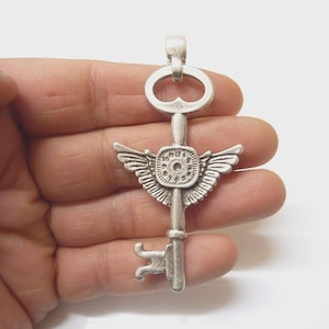 Può includere: Un ciondolo a forma di chiave color argento con un design dettagliato. La chiave presenta un elemento circolare con ali e un quadrante dell'orologio, e una forma di buco della serratura nella parte inferiore. Il ciondolo è presentato su sfondo bianco.