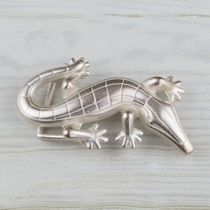 May include: Silver-toned alligator belt buckle with a detailed, textured design. The buckle is shaped like an alligator with its mouth open.
