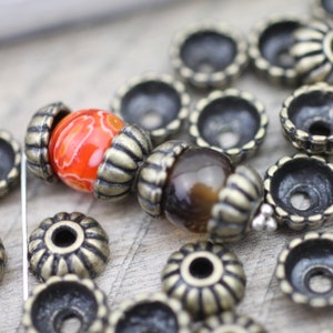 May include: A close-up of two beads with ornate metal caps. The beads are orange and brown with a swirl pattern. The beads are surrounded by many small, round, metal caps.