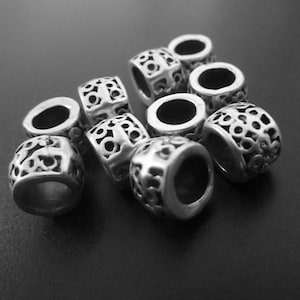 Puede incluir: Un grupo de cuentas de metal plateadas con diseños intrincados. Las cuentas son redondas y están diseñadas para usarse como separadores o dijes en un brazalete o collar.