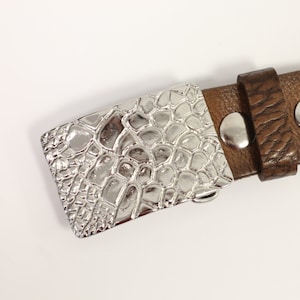 Pode incluir: Uma fivela de cinto prateada com um padrão de pele de crocodilo. A fivela está presa a um cinto de couro castanho.