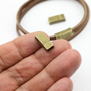 May include: A close-up of antique brass magnetic clasps for jewelry making. The rectangular clasps are shown with a brown leather bracelet in the background. The clasps are designed to connect and secure the bracelet.