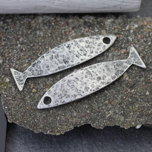 May include: Two silver fish charms with a hammered texture. The charms have a hole at the top for attaching to a necklace or bracelet.
