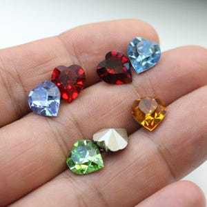 Peut inclure: Assortiment de six strass en forme de cœur de différentes couleurs, dont le rouge, le bleu, le vert et l'or. Les strass sont présentés sur une main.