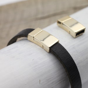 Pode incluir: Um bracelete de couro preto com um fecho magnético dourado. O fecho é mostrado fechado e aberto.