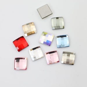 Puede incluir: Una colección de pedrería cuadrada y facetada en varios colores, incluyendo rojo, rosa, dorado e iridiscente. La pedrería está dispersa sobre una superficie blanca, mostrando sus cualidades reflectantes y formas geométricas.