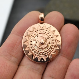 May include: A rose gold pendant featuring a circular design with a starburst pattern and a concentric circle design in the center.