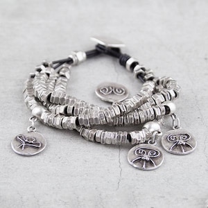 Può includere: Un braccialetto d'argento con più file di perline e tre ciondoli d'argento. I ciondoli sono circolari e presentano un design stilizzato.