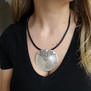 May include: A silver pendant necklace featuring a large, circular pendant with an ornate top section. The pendant hangs from a black cord. The necklace is worn against a black top.