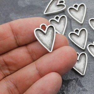 May include: A close-up of a hand holding a silver heart-shaped charm. The charm has a slightly textured surface and a hollow center. There are several other similar charms in the background.