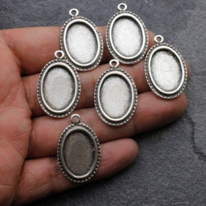 May include: Six silver oval-shaped pendant trays with a beaded edge. The trays are blank and ready to be filled with resin or other materials.