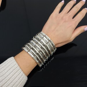 May include: A silver bracelet with a stacked design. The bracelet is made of multiple rows of connected, textured, rectangular pieces.
