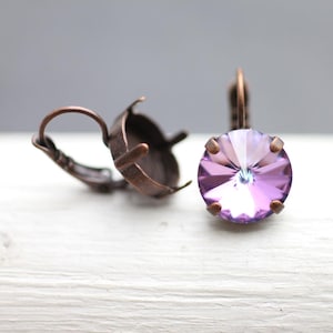 Pode incluir: Um par de engastes de brincos em tom cobre com um único cristal roxo em um engaste. O cristal tem um design facetado e um tom roxo claro.