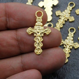 May include: Gold-toned metal cross charms with a detailed design of a figure on the cross. The charms have a loop at the top for attaching to jewelry or other crafts.