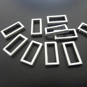 Puede incluir: Un grupo de abalorios rectangulares de metal plateado con dos pequeños agujeros a cada lado. Los abalorios están esparcidos sobre una superficie negra.