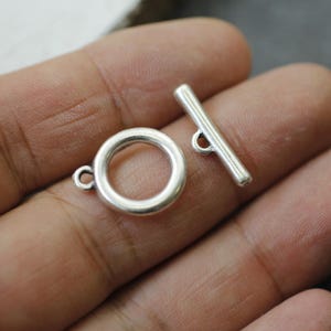 Op de afbeelding: Een zilverkleurige toggle sluiting set, bestaande uit een cirkelvormige ring met een kleine lus en een rechte staaf met een lus. De sluiting is ontworpen voor het maken van sieraden en knutselen. Het metaal heeft een glanzende afwerking.