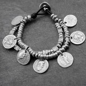 May include: A silver bracelet with a leather cord and multiple coin charms. The coins have various designs and are attached to the bracelet with small silver rings.