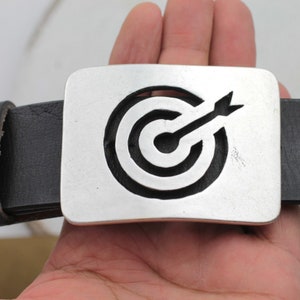 May include: A silver belt buckle with a black leather belt. The buckle features a stylized target with an arrow in the center.