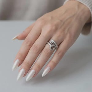 Pode incluir: Um anel de metal em tom prateado com um design envolvente em forma de chave antiga. O anel tem um acabamento fosco e é usado no dedo anelar, exibindo a cabeça da chave e a haste que envolve a banda do anel.